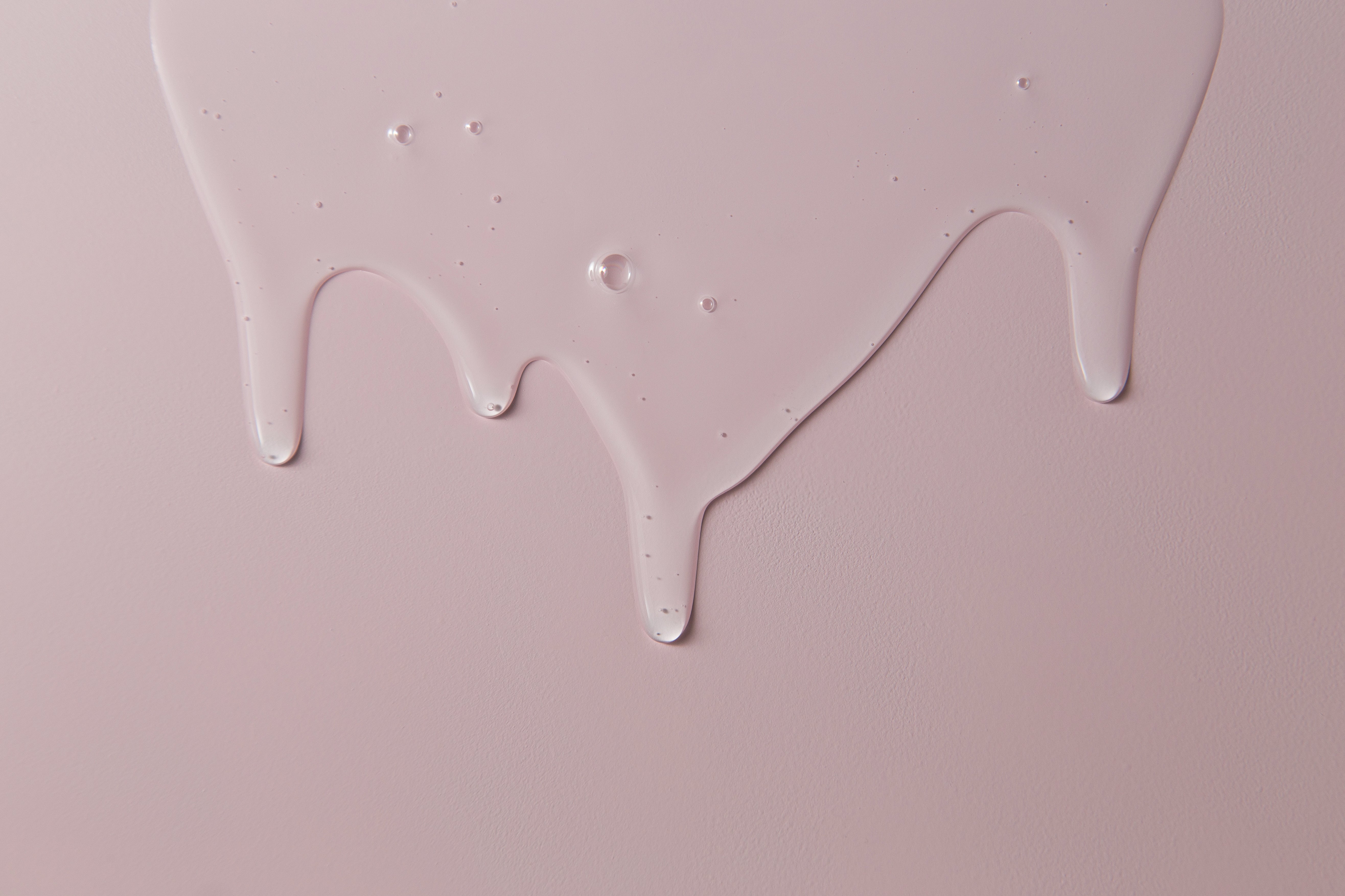 A smooth, light pink liquid like Dr Loretta — Minis drips down a pastel pink surface, creating rounded droplets and glossy trails.