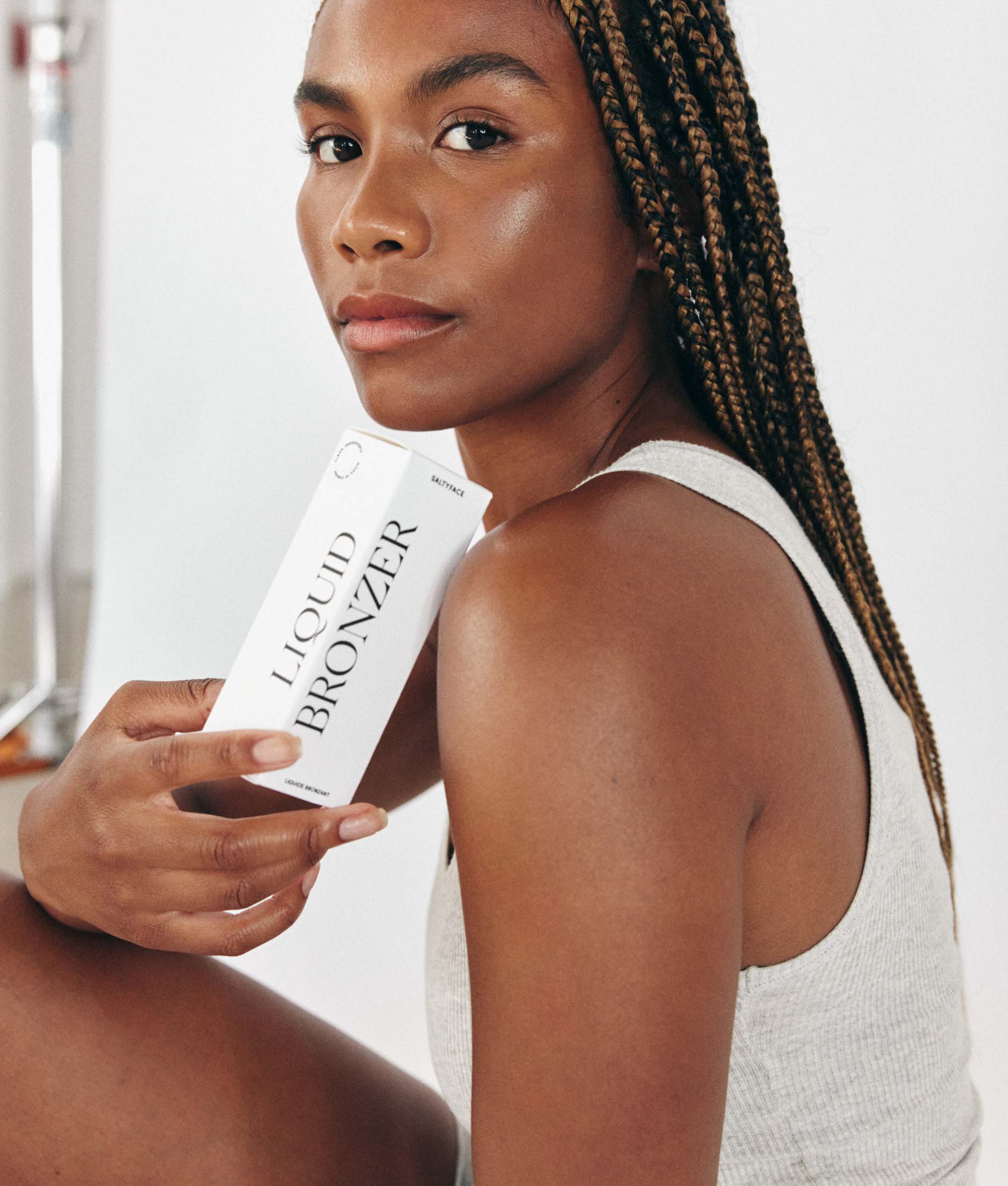 A model in a grey tank looking at the camera and holding a box of Liquid Bronzer. 