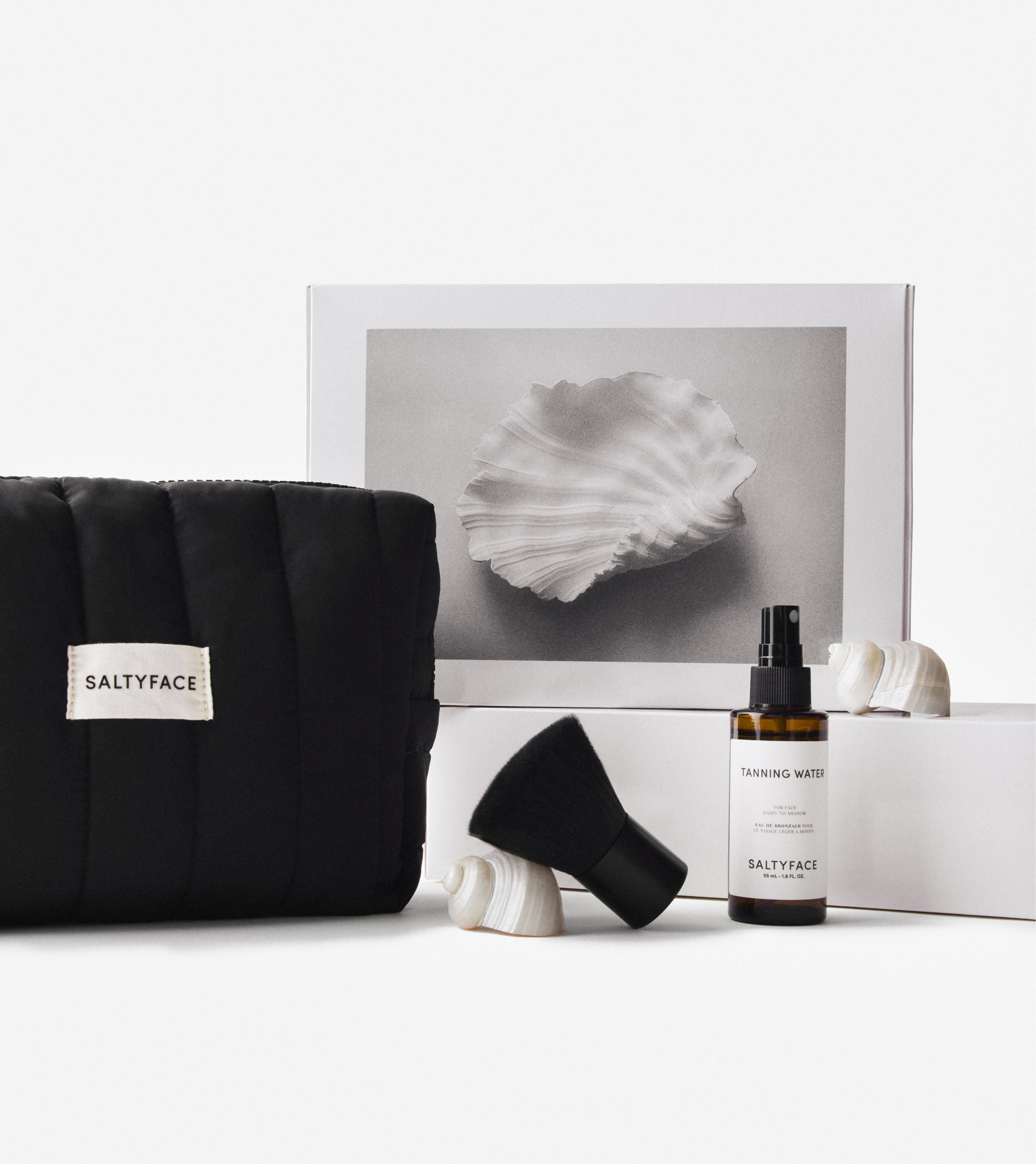 Black The Everywhere travel bag with a Saltyface logo, Brush No. 1 and Saltyface Tanning Water are displayed with a white gift box with a seashell on it.